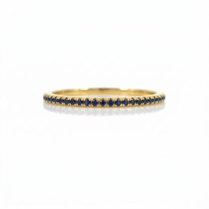 A thin yellow gold eternity ring set with a single row of small round dark blue sapphires, shown on a clean white background with a soft reflection.