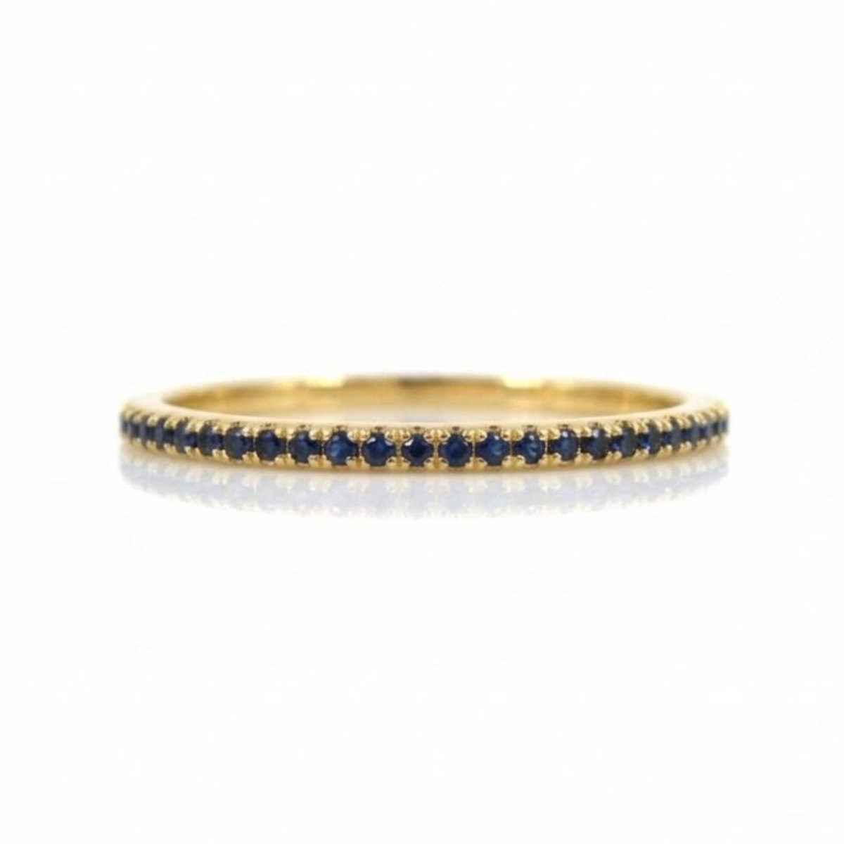 A thin yellow gold eternity ring set with a single row of round-cut deep blue sapphires, shown on a clean white background with a soft reflection.