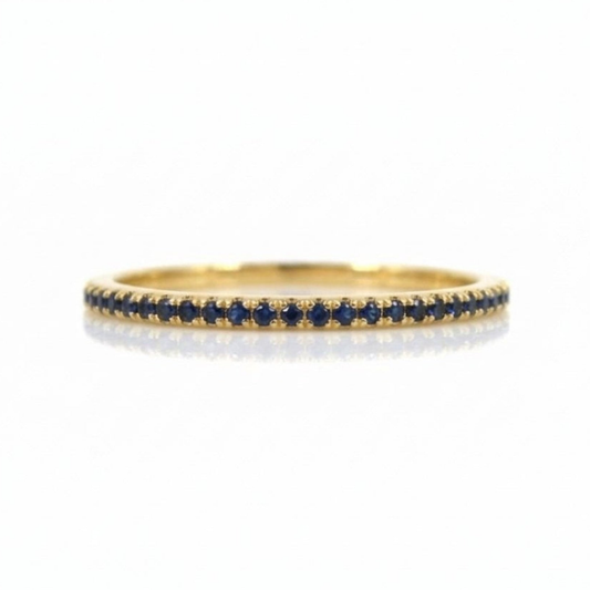 A thin yellow gold eternity ring set with a single row of round-cut deep blue sapphires, shown on a clean white background with a soft reflection.