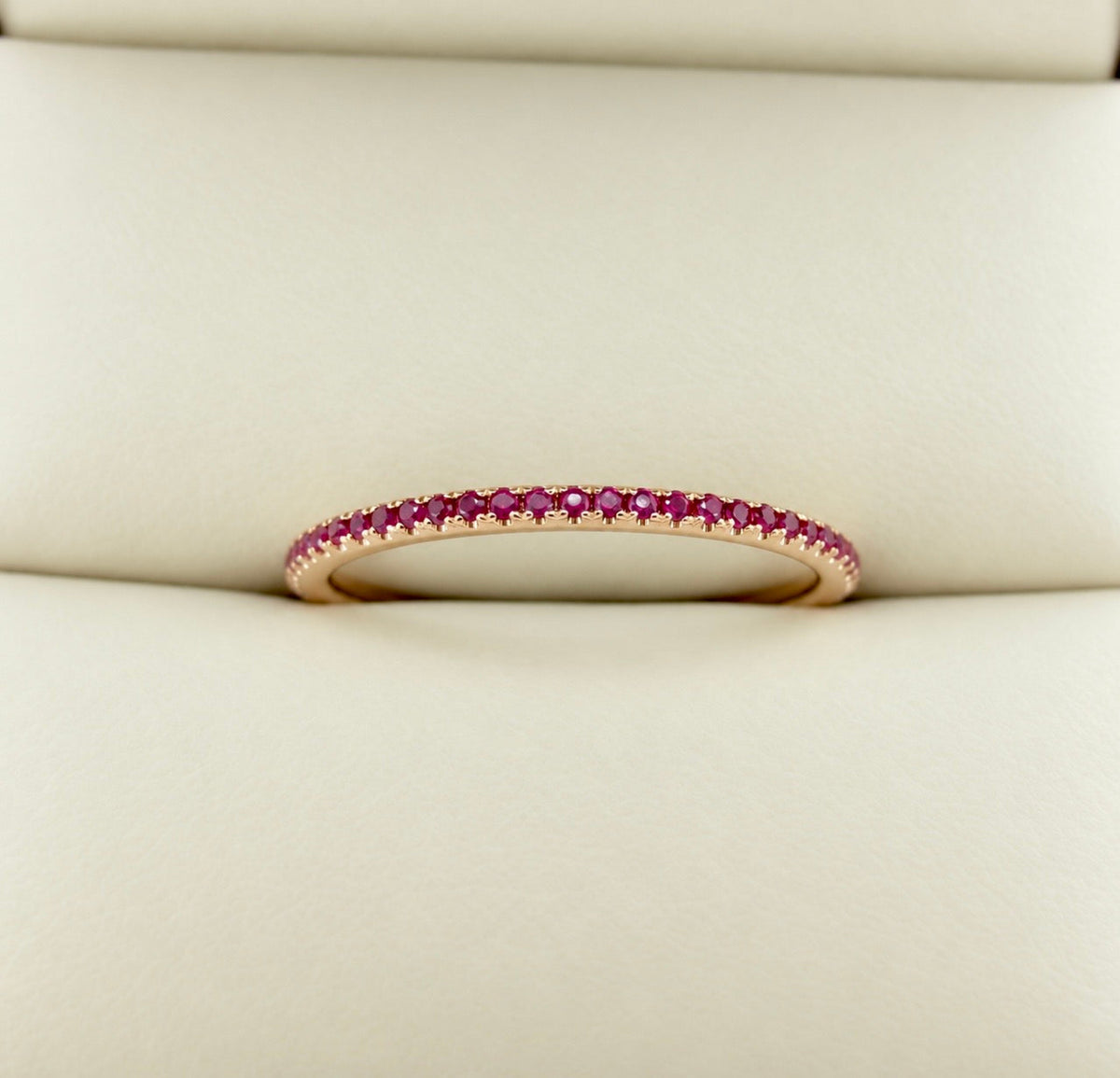 A delicate rose gold eternity ring set with small round red rubies, resting on a cream-colored textured surface.