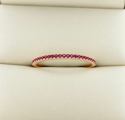 A delicate rose gold eternity ring set with small round red rubies, resting on a cream-colored textured surface.