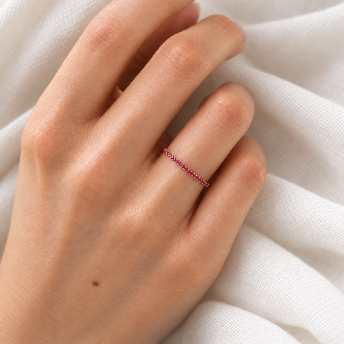 A close-up of a hand wearing a delicate ruby ring, resting against soft white fabric.