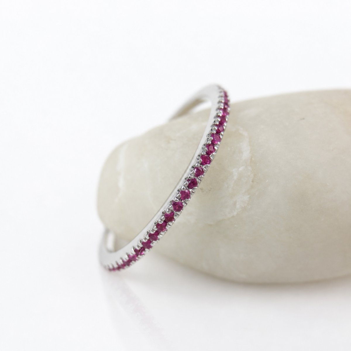 A thin silver ring set with a row of small red rubies, displayed on a smooth white stone against a clean white background.