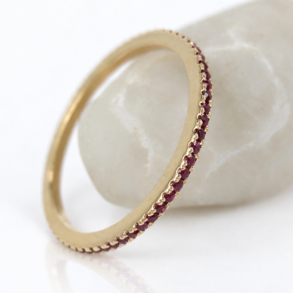 A gold eternity ring with a single row of small round red rubies, displayed on a smooth white stone prop against a white background.
