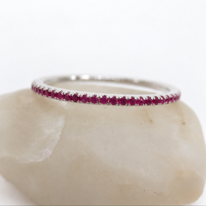 A thin silver band ring set with a row of small round red rubies, displayed on a smooth light beige stone against a white background.