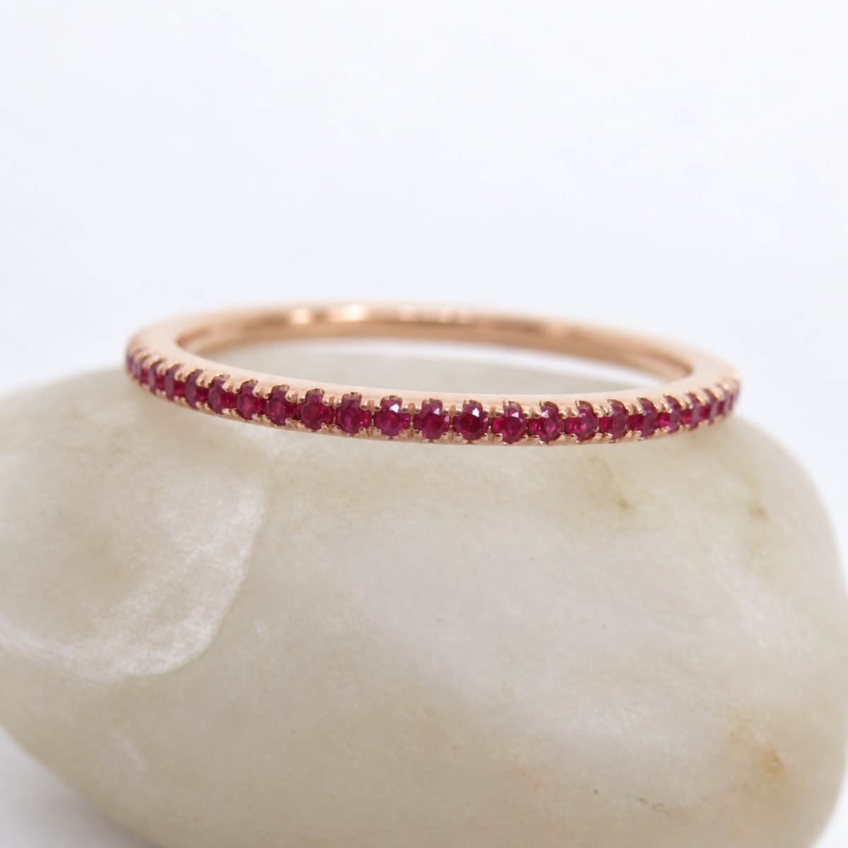 A thin rose gold eternity ring set with small round red rubies, resting on a smooth white stone against a white background.